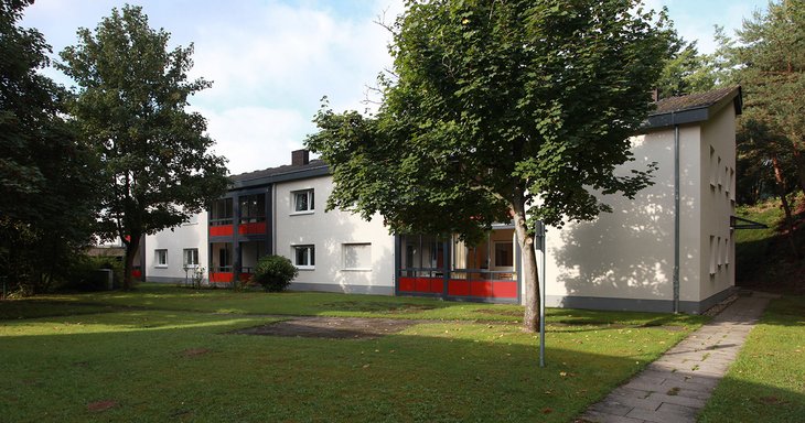 Hier ist das sogenannte Teilhabezentrum in Dahn, ein Wohnangebot des Pfalzklinikums zu sehen. Das Gebäude hell und rötlich gestrichen und sieht sehr modern aus. Neben dem Gebäude sind viele Bäume und eine Wiese zu sehen.