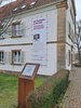 Zu sehen ist das Alleehaus mit einem Werbebanner für die Ausstellung und einer Informationsstele.