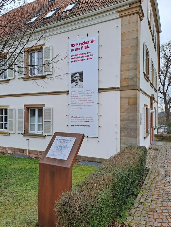 Zu sehen ist das Alleehaus mit einem Werbebanner für die Ausstellung und einer Informationsstele.