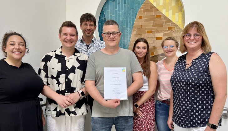 Auf dem Bild sind 7 Mitarbeitende des Pfalzklinikums zu sehen, die stolz die Urkunde zum gewonnen Preis in die Kamera halten.