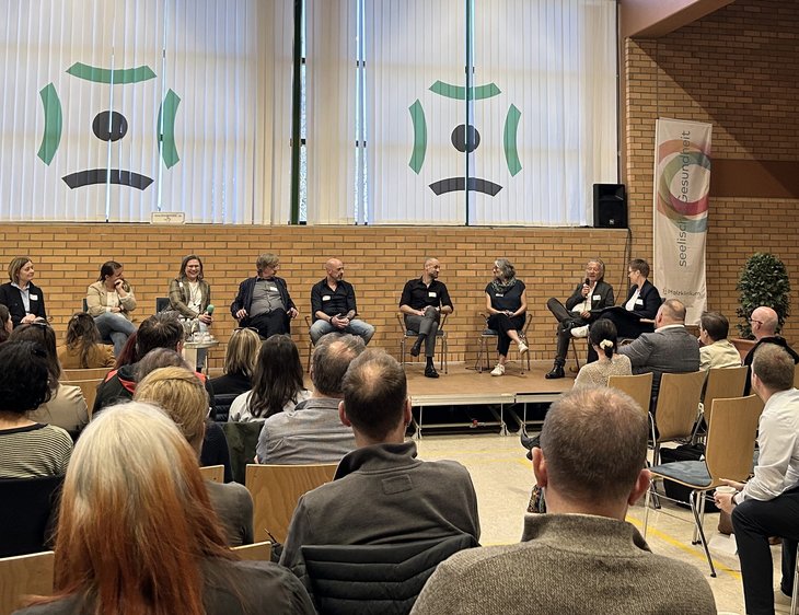 Podiumsdiskussion auf dem Symposium. 9 Menschen sitzen im Halbkreis in einem Gespräch auf einer Bühne.