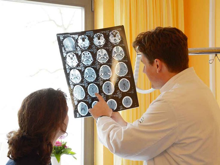 Ein Arzt ist im Gespräch mit einer Patientin in einem freundlich gestalteten Zimmer der Klinik für Neurologie. Er zeigt ihr CT oder Röntgenaufnahmen und erklärt ihr die Befunde der Untersuchung. Die Patientin ist nur von hinten zu sehen, der Arzt ebenfalls.