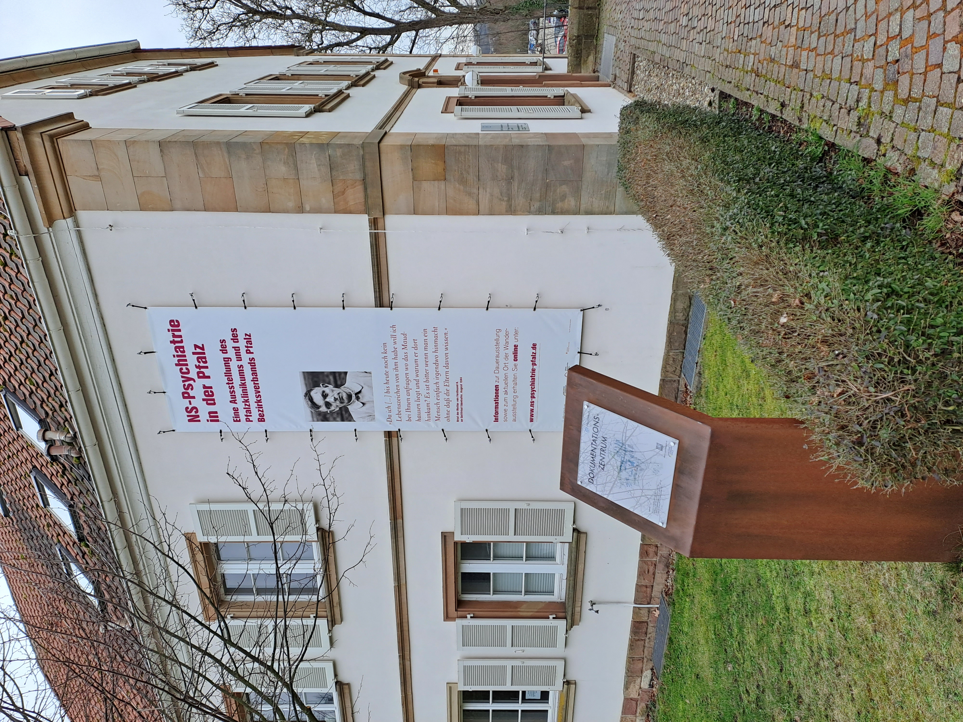 Zu sehen ist das Alleehaus mit einem Werbebanner für die Ausstellung und einer Informationsstele.